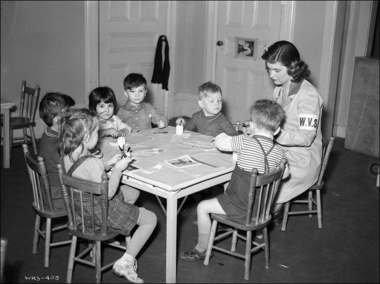 Une assistante du Service féminin bénévole aide les enfants à faire un collage d’un hors-texte dans une garderie de jour de l’école Givens, Toronto (Ontario), février 1943